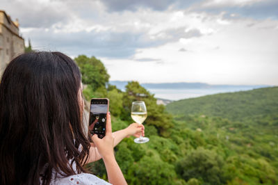 Rear view of young woman taking photos of a glass of wine in front of beautiful landscape view