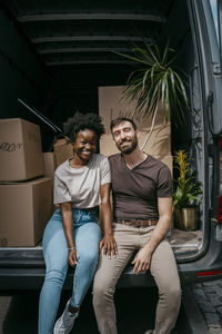 Portrait of happy boyfriend and girlfriend sitting together in van trunk with boxes