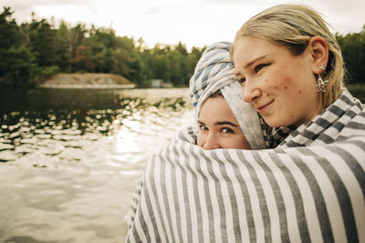 Woman embracing female friend wrapped in towel at lake during vacation