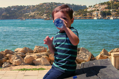 Boy drinking water from bottle while sitting against sea