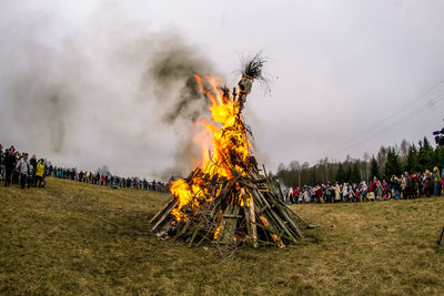 Crowd gathered by scarecrow burning on field against sky