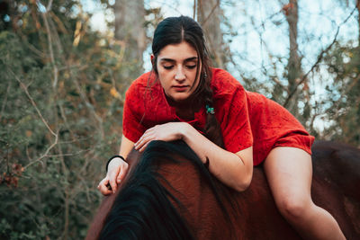 Beautiful young woman lying down on horse against tree