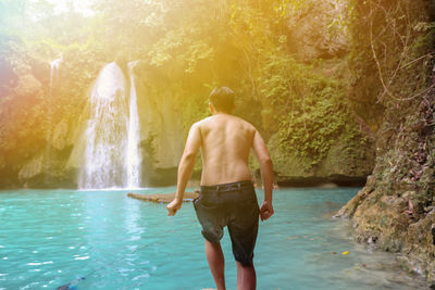 Rear view of shirtless man standing against waterfall