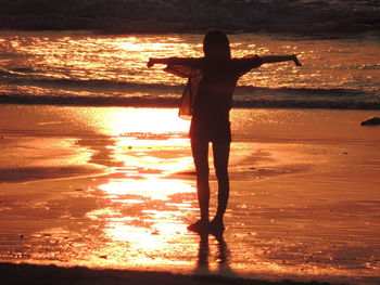 Rear view of woman with arms outstretched standing on beach during sunset