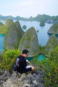 Rear view of asian man sitting on rocky cliff by the sea of islands in raja ampat, papua, indonesia