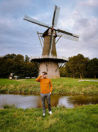 Full length of man wearing hat standing by pond against windmill on field