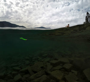 Water surface shot of boy fishing in river against cloudy sky