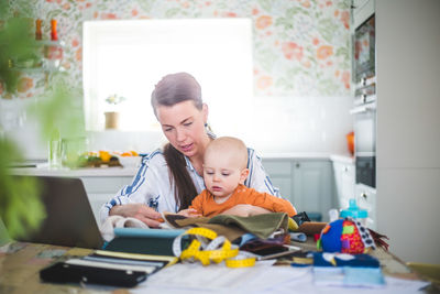 Fashion designer taking care of daughter while working at table in kitchen at home