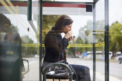 Side view of businesswoman eating sandwich while waiting at bus stop in city