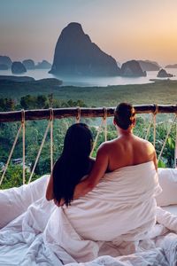 Rear view of couple covered under blanket while sitting against mountain during sunset
