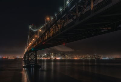 Low angle view of illuminated bay bridge over river against sky at night