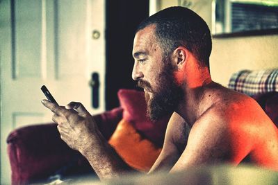 Shirtless bearded man using phone while sitting on sofa at home