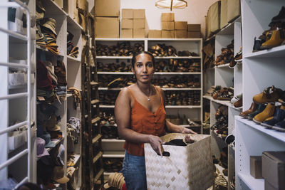 Female clerk carrying wicker basket while standing amidst shoe racks at store