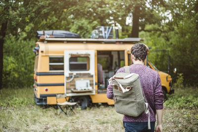 Rear view of young man walking with backpack towards motor home in forest during camping