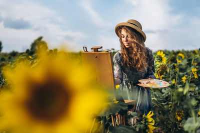 A young woman with curly hair and wearing a hat is painting in nature. 