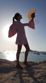 Full length of young woman with hand fan standing on seashore against clear sky