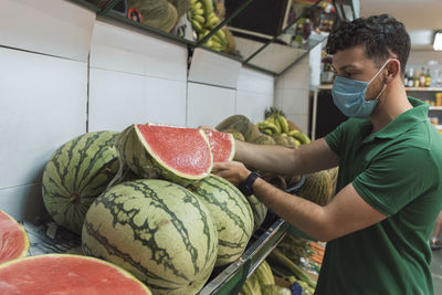 A young man wearing a mask placing watermelons in the fruit shop.