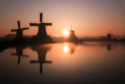 Silhouette traditional windmills by lake against clear sky during sunset