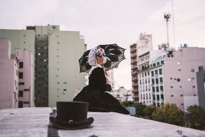 Woman with umbrella sitting on rooftop against sky in city