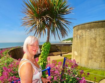 Portrait of smiling mature woman standing by plants against blue sky during sunny day