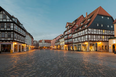 Illuminated street amidst buildings against sky at dusk