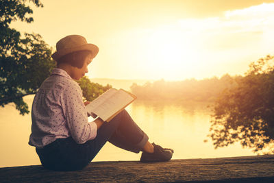 Man sitting by lake against sky during sunset