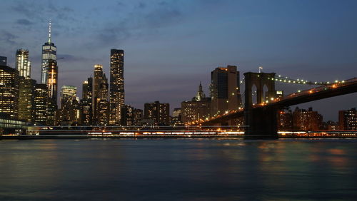 Illuminated bridge over river by buildings against sky at night