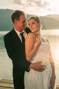 Smiling bridegroom holding bride while standing on pier against lake