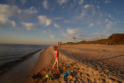 Girl with arms raised standing at beach against sky during sunset