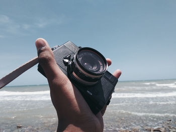 Midsection of person holding camera at beach against sky