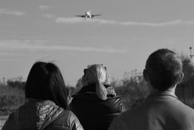 Rear view of people watching flying airplane against sky