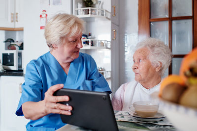 Caring nurse in uniform demonstrating information on modern tablet while sitting at table with meal in light kitchen at home