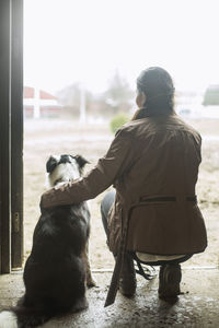 Full length rear view of young woman arm around dog in doorway of horse stable