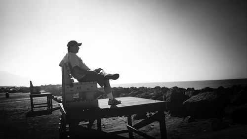 Side view of man relaxing on wooden bench by sea against sky