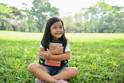 Smiling girl with books looking away while sitting on grassy field at park