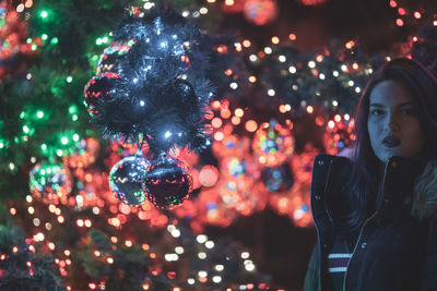 Portrait of young woman standing against illuminated christmas tree at night
