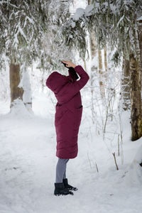 A girl in a burgundy down jacket photographs a snow-covered forest on a mobile phone. person