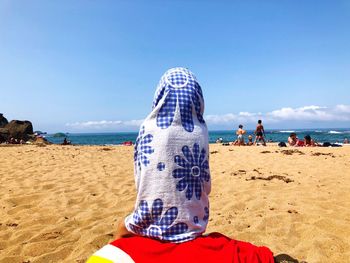 Rear view of person wrapped in towel sitting at beach against sky