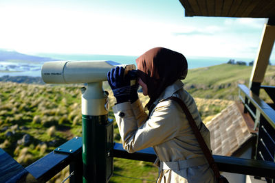 Side view of woman looking through coin-operated binoculars against sky