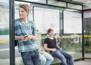 Young man holding mobile phone while waiting with friends at bus stop