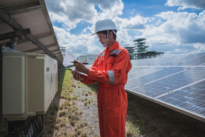 Side view of worker wearing reflective clothing while standing on solar panel against sky