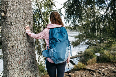 A girl with a backpack walks in the spring forest, put her hand on a tree and admires the view.