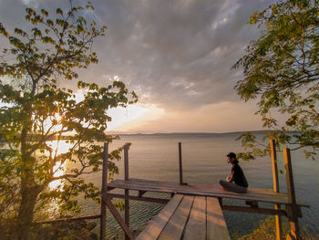 Woman sitting on railing by sea against sky