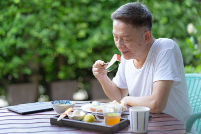 Smiling senior man eating watermelon while sitting on chair outdoors