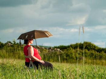 Woman with umbrella sitting on field against sky