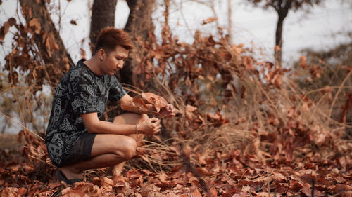 Side view of young man crouching while holding dry leaf