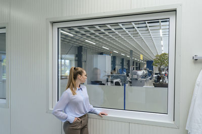 Young businesswoman with hand in pocket looking through window at warehouse