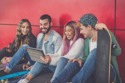 Full length of cheerful friends looking at digital tablet while sitting against red wall