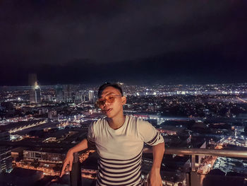 Portrait of man standing against illuminated cityscape at night