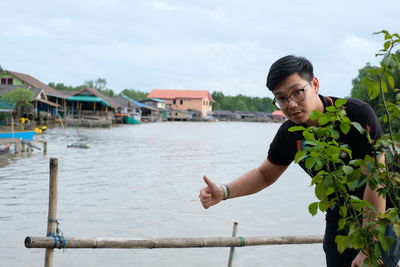 Portrait of young man wearing eyeglasses standing by lake against cloudy sky